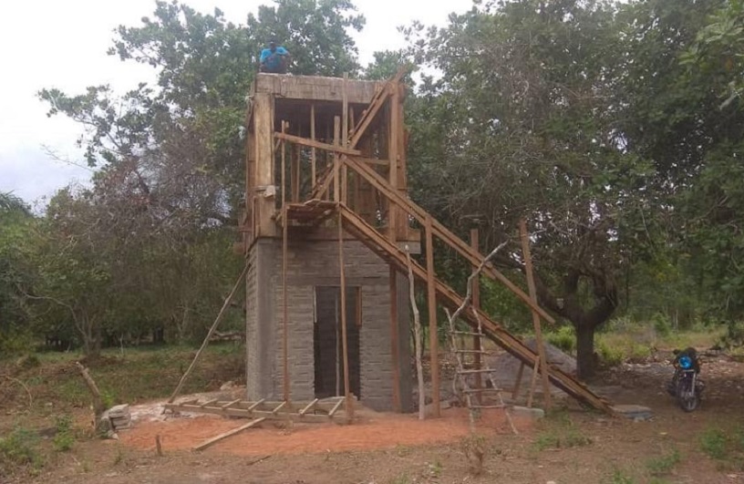 Ein unvollendeter Bau mit Holzrahmen und Treppe, umgeben von Bäumen und Gras. Eine Person arbeitet auf dem Dach.