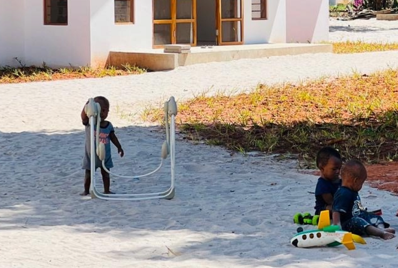 Zwei Kinder spielen auf dem Boden vor einem weißen Gebäude, während ein weiteres Kind an einem Spielzeug zieht.