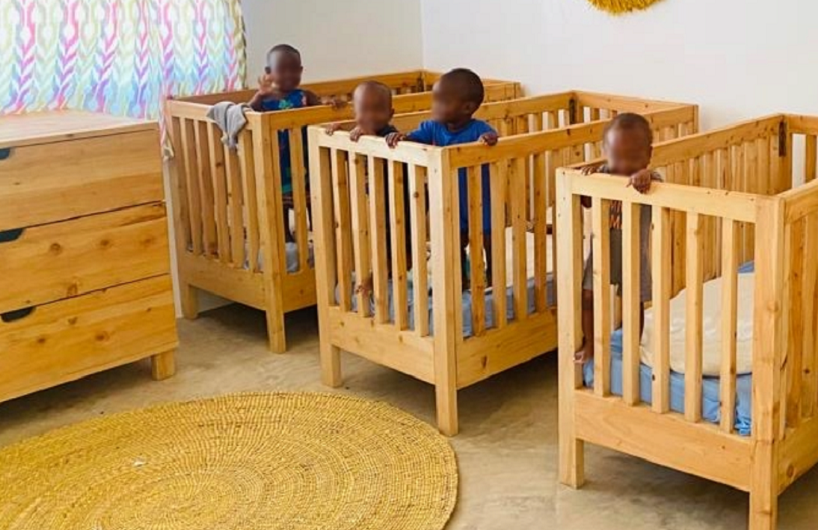 Drei Kinder stehen in Holz-Kinderbetten in einem hellen Raum mit bunten Vorhängen und einem runden Teppich.