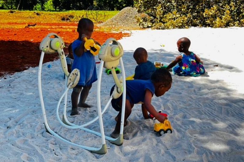 Kinder spielen im Sand, während ein weiteres Kind mit einem Spielzeug beschäftigt ist. Die Umgebung zeigt viel Grün und einen klaren Himmel.
