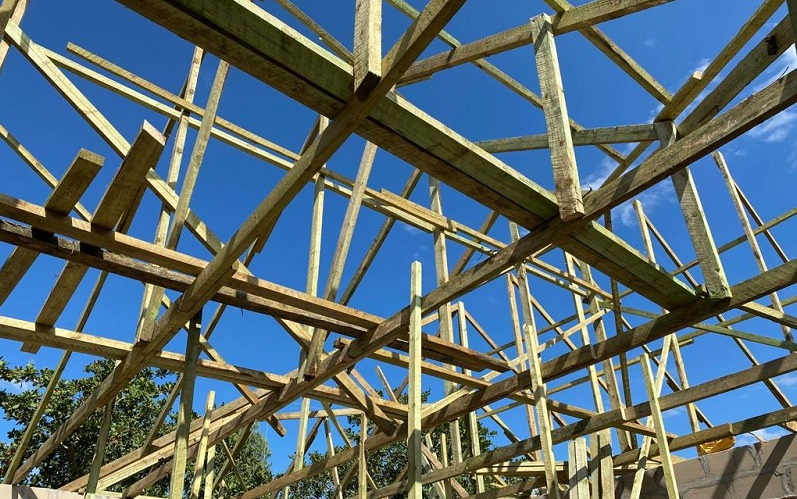 Holzrahmenkonstruktion unter blauem Himmel, mit sichtbaren Balken und Stützen, die den Bau eines Gebäudes unterstützen.