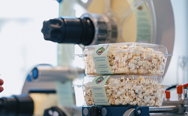 Zwei durchsichtige Behälter mit Popcorn stehen übereinander, während im Hintergrund eine Maschine zur Popcornherstellung sichtbar ist.