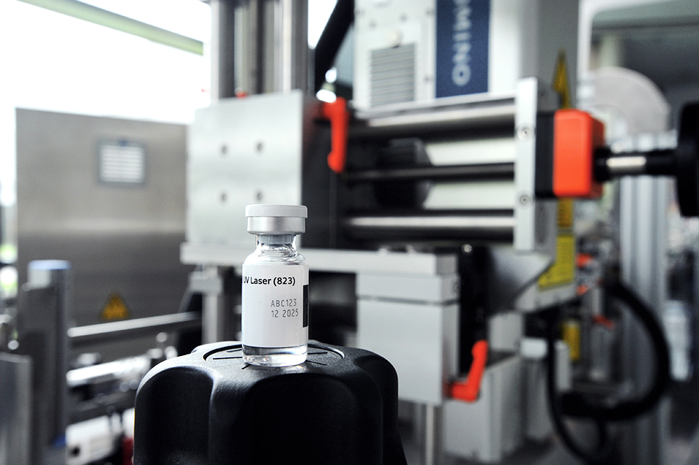 Eine Spritze mit einer klaren Flüssigkeit steht auf einem schwarzen Deckel vor einer industriellen Maschine.