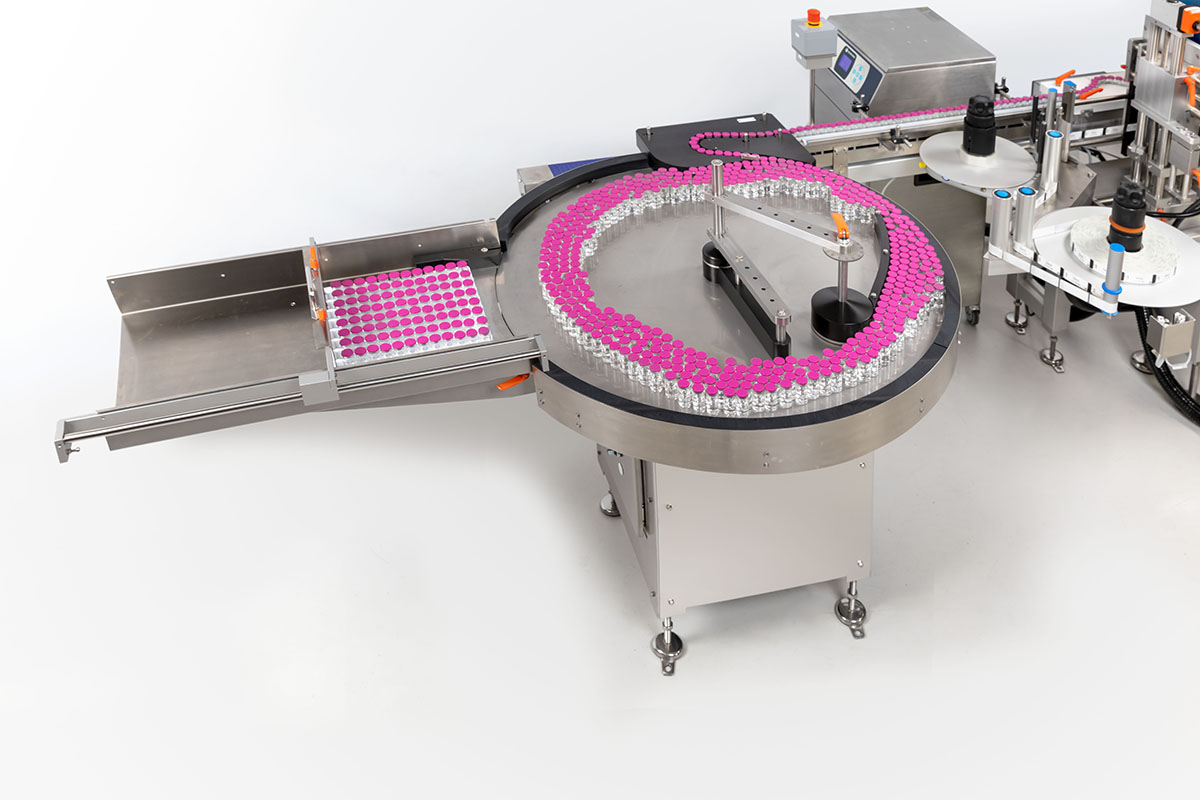 Automatisierte Abfüllanlage mit runder Förderstrecke, die mit kleinen Fläschchen gefüllt ist. Pinke Etiketten sind sichtbar.