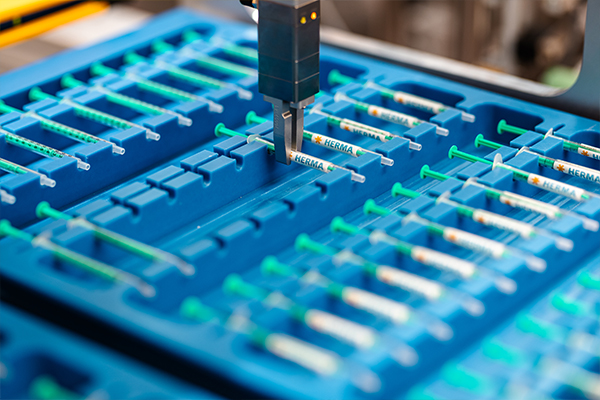 Ein automatisiertes Gerät entnimmt Spritzen aus einem blauen Behälter mit mehreren Fächern, die für die Lagerung von Spritzen ausgelegt sind.