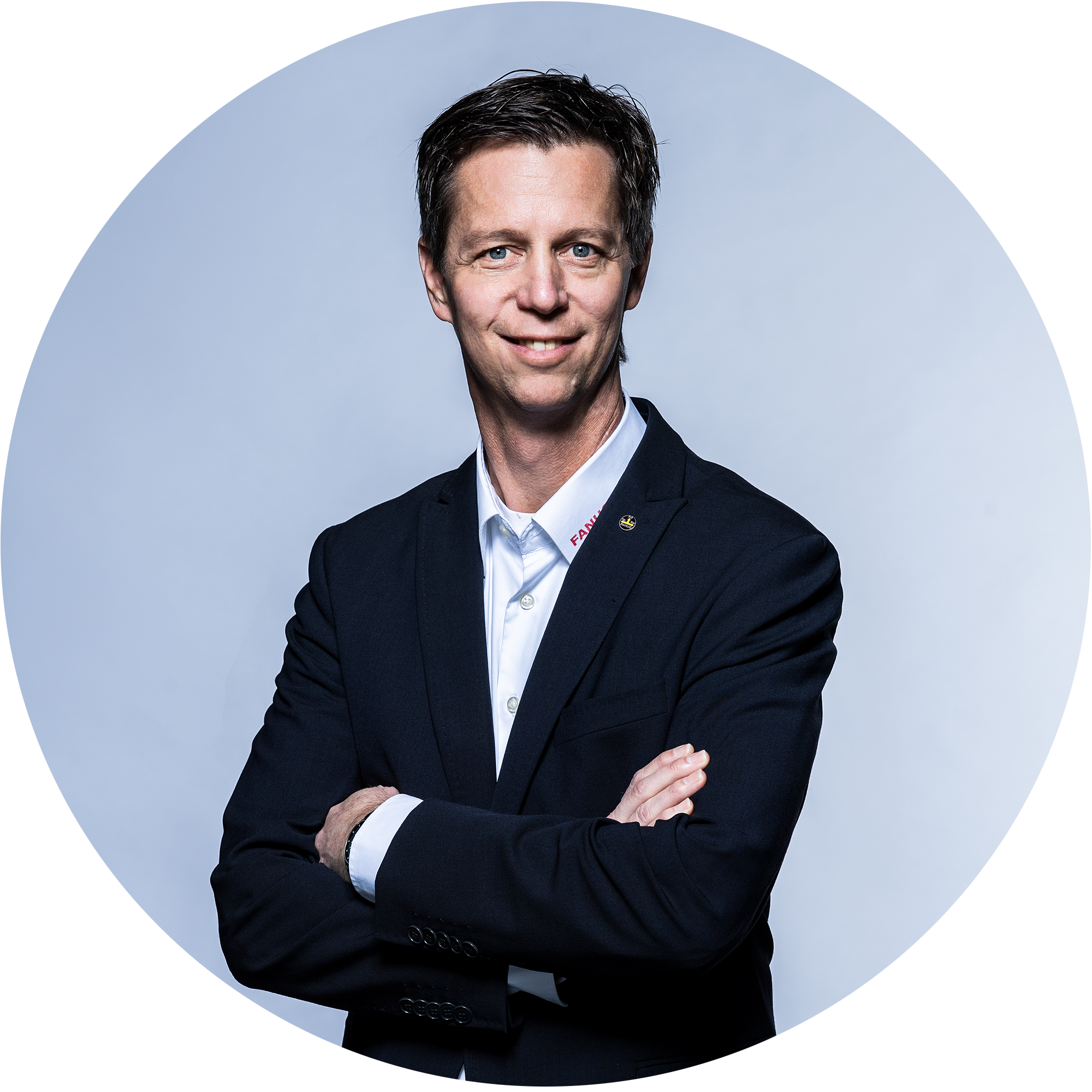 Ein Mann in einem schwarzen Anzug mit verschränkten Armen lächelt in die Kamera. Der Hintergrund ist hell und neutral.