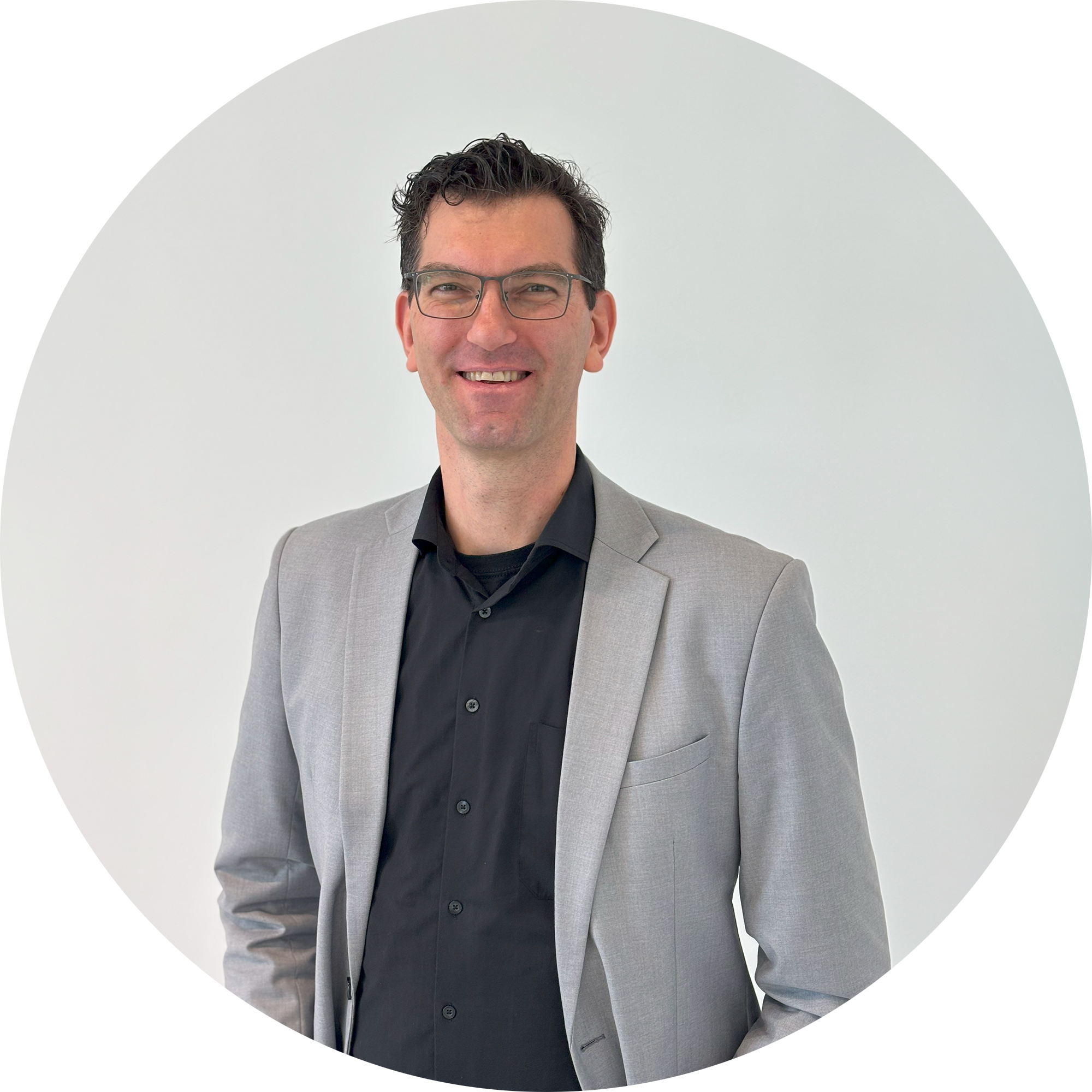 Ein Mann mit Brille trägt einen grauen Blazer über einem schwarzen Hemd und lächelt vor einem hellen Hintergrund.