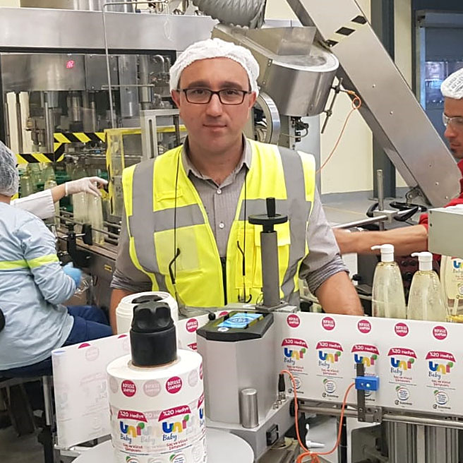 Mann in Sicherheitskleidung und Haarnetz steht in einer Produktionsstätte, während Mitarbeiter Flaschen abfüllen. Maschinen im Hintergrund.