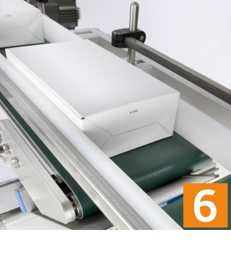 Ein Förderband transportiert mehrere weiße Verpackungen, während eine grüne Unterlage sichtbar ist. Eine orange Nummer 6 ist in der Ecke zu sehen.
