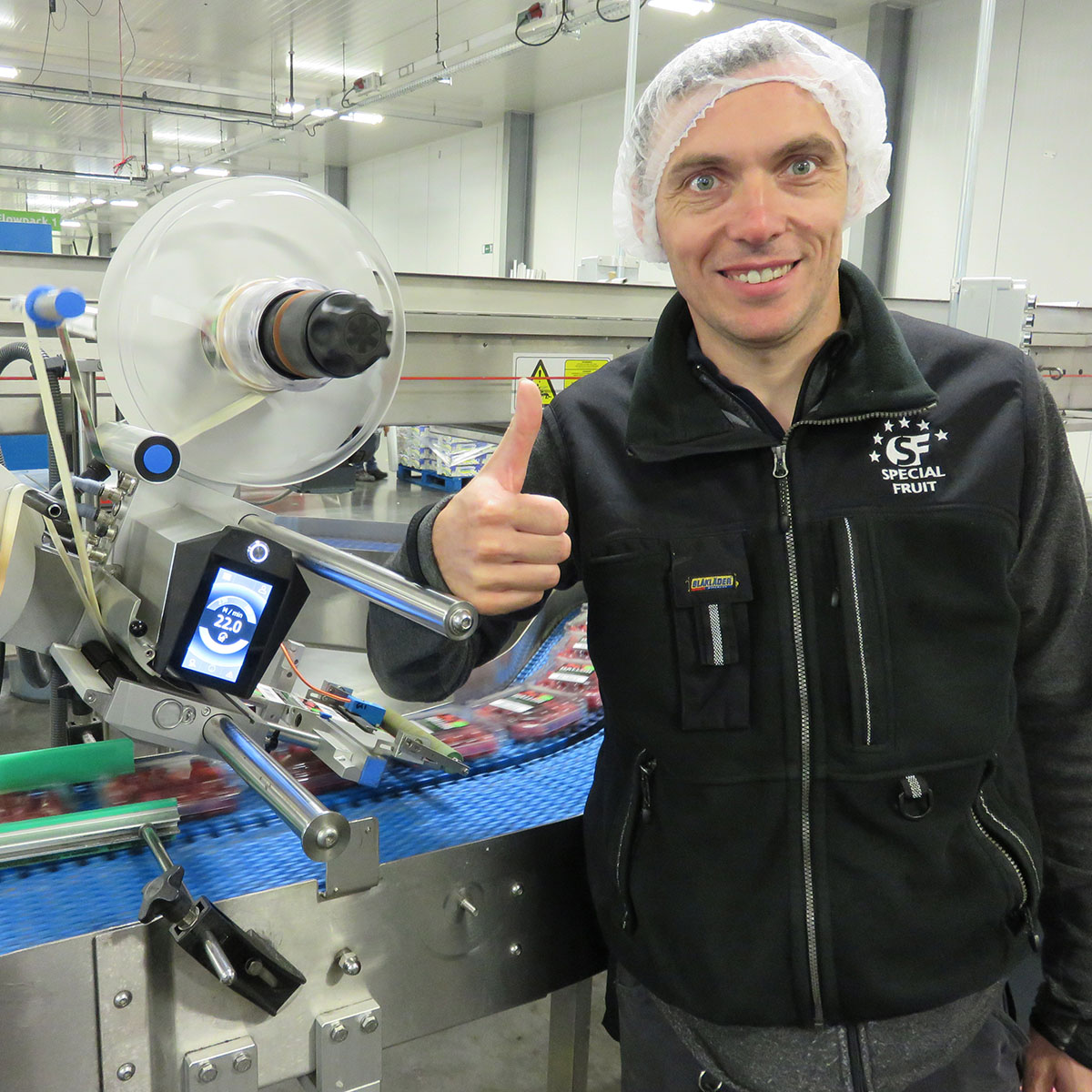Mann mit Daumen hoch in einer Produktionsstätte, neben einer Maschine zur Etikettierung von Produkten auf einem Förderband.