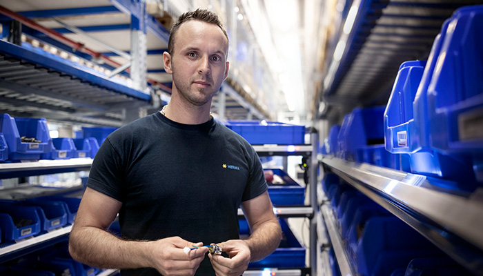 Ein Mann in einem schwarzen T-Shirt hält ein Werkzeug in der Hand, umgeben von blauen Aufbewahrungsboxen in einem Lagerregal.