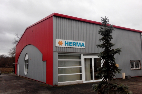 Fassade eines modernen Gebäudes mit rotem Dach und grauen Wänden, beschriftet mit "HERMA". Ein Baum steht im Vordergrund.