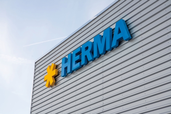 Das Firmenlogo von HERMA in blauer Schrift mit einem gelben Stern auf einer grauen Wand.