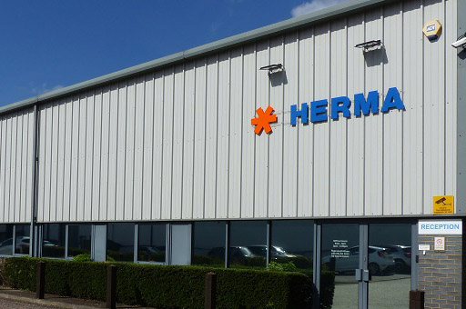 Fassade eines modernen Gebäudes mit dem Schriftzug "HERMA" in blauer Schrift und einem orangefarbenen Symbol. Empfangsbereich sichtbar.