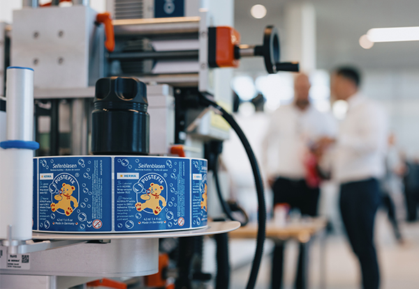 Eine Verpackungsmaschine mit bunten Etiketten auf Dosen im Vordergrund, während Personen im Hintergrund miteinander sprechen.