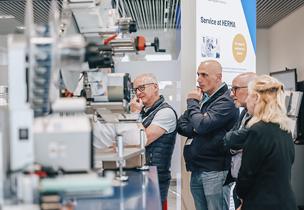 Vier Personen beobachten aufmerksam eine Maschinenpräsentation auf einer Messe, während Informationen über den Service bei HERMA angezeigt werden.