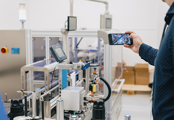 Eine Person hält ein Smartphone und filmt eine automatisierte Maschinenanlage in einer Produktionsumgebung.