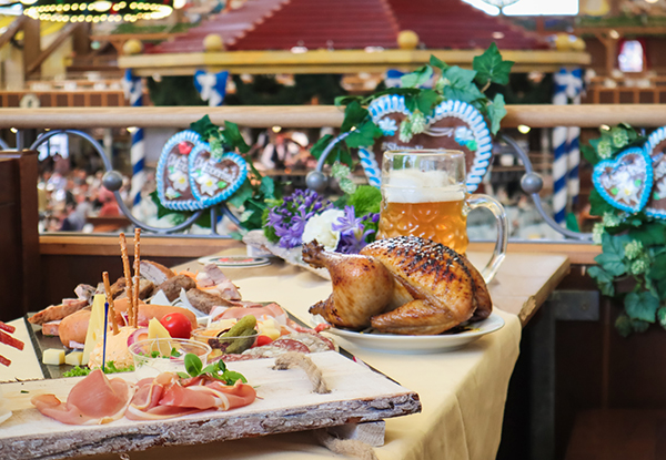 Ein Tisch mit einer Auswahl an Wurst, Käse und Gemüse, dazu ein gebratenes Hähnchen und ein großes Bierglas in einem festlichen Ambiente.