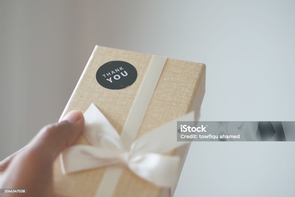 Eine Person hält ein Geschenk in der Hand, das mit einem weißen Band und einem schwarzen "THANK YOU"-Aufkleber verziert ist.