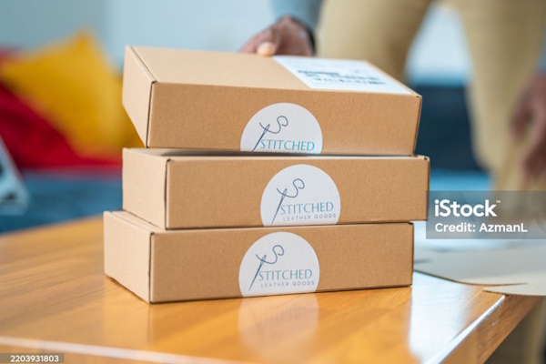 Drei braune Kartons mit dem Aufdruck "Stitched" stehen auf einem Tisch, während eine Person im Hintergrund eine Hand darauf legt.