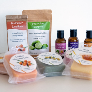 Etiketten auf verschiedenen Kosmetikprodukten: Doypacks mit Bademilch und Badepulver, Braunglasflaschen mit Badearoma sowie Pflanzenöl-Seifen mit runden Etiketten in Klarsichtbeuteln, auf hellem Hintergrund.