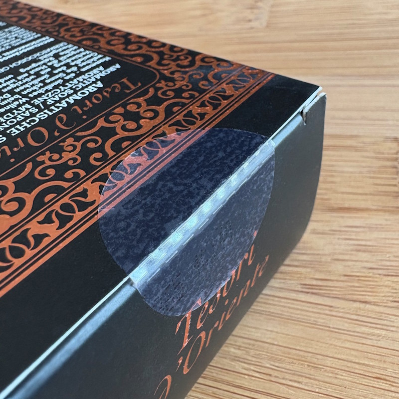 Lösungen Kosmetik: Schwarze Verpackung mit dekorativen, orangen Mustern und Text. Die Lasche der Verpackung ist mit einem transparenten Verschlussetikett versiegelt