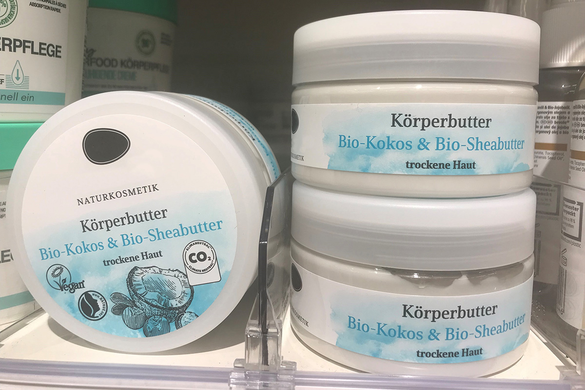 Weiße Cremedose mit Naturkosmetik-Körperbutter im Regal, etikettiert auf Deckel und Korpus mit farbig bedruckten Etiketten in Blau-Weiß.