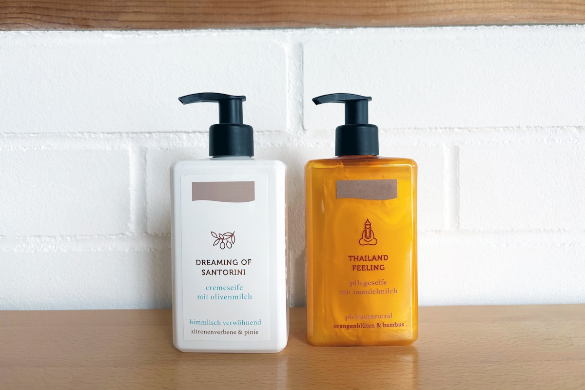 Zwei Pumpspender-Flaschen mit Cremeseife: eine weiße Flasche mit weißem Etikett und eine orangefarbene Flasche mit transparentem Folienetikett im No-Label-Look, auf einer Holzoberfläche vor weißer Ziegelwand.