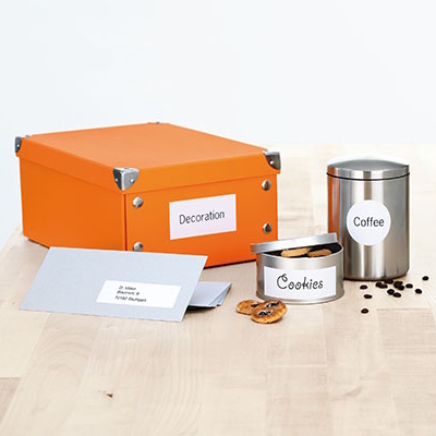 Bedruckte Universaletiketten auf orangener Box, silbernem Kaffeebehälter, Dose mit Cookies und grauem Umschlag auf einem Holztisch.