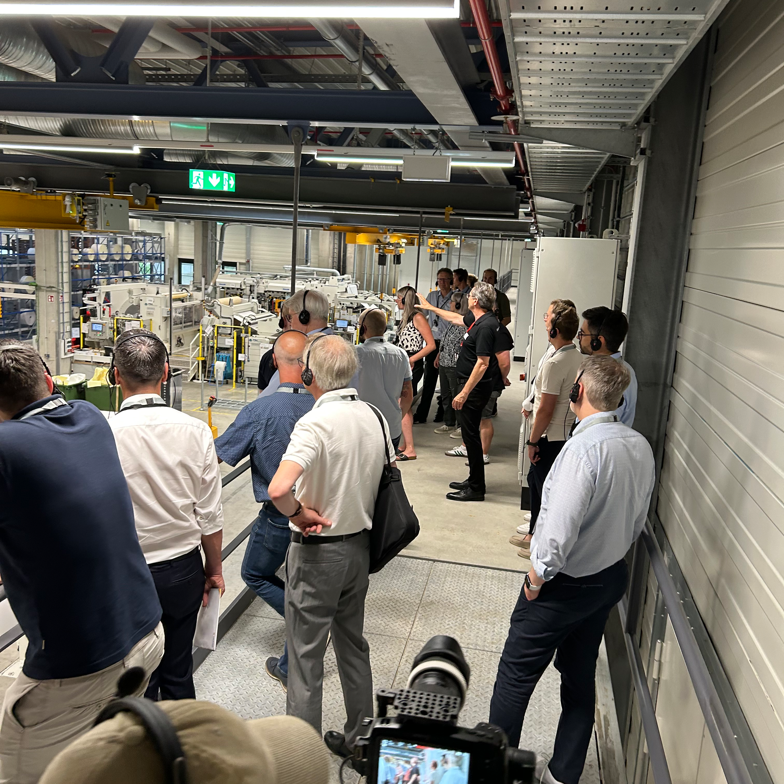 Gruppenschulung in einer Produktionsstätte, Teilnehmer beobachten Maschinen und diskutieren. Einige Personen machen Notizen oder fotografieren.