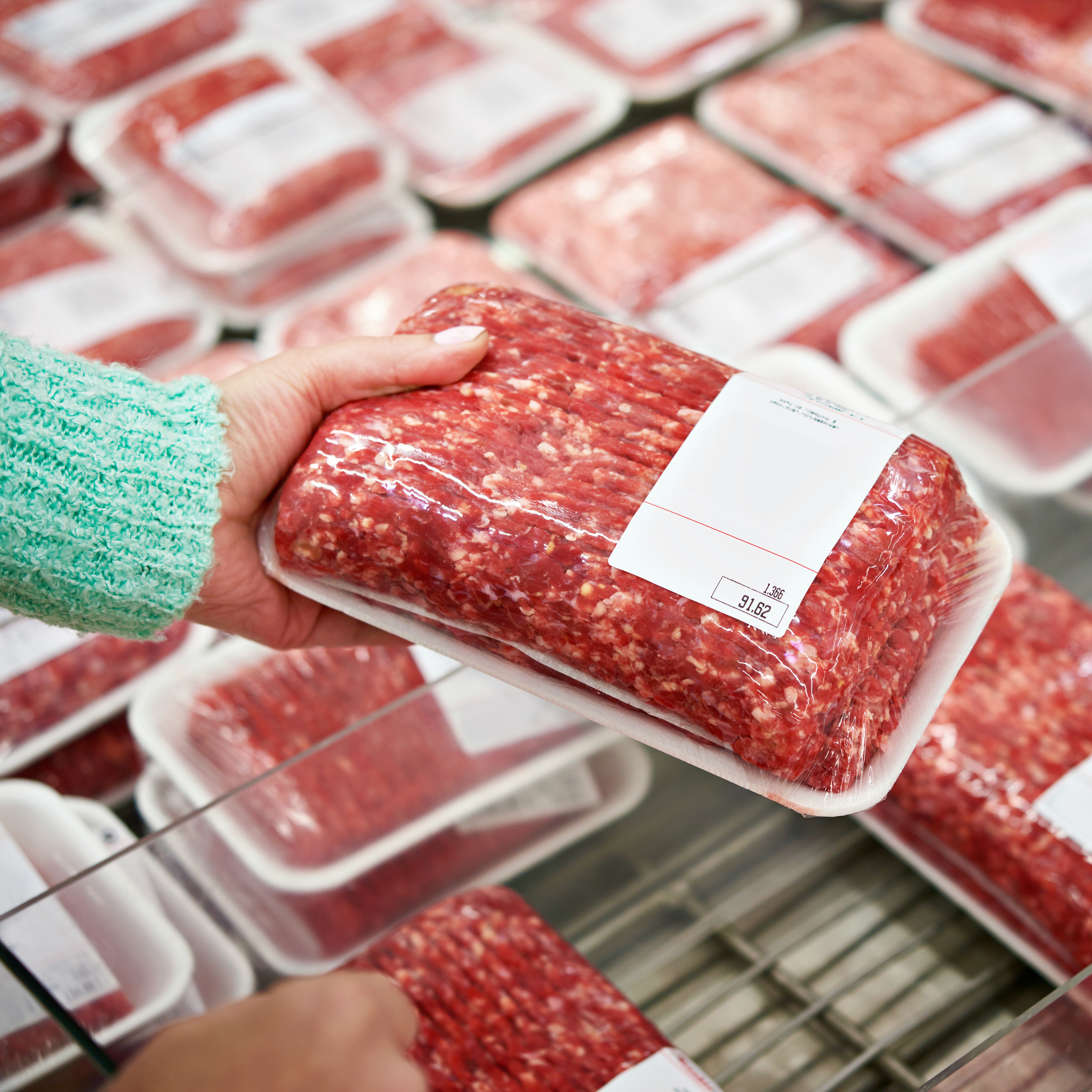 Eine Hand hält ein Paket mit Hackfleisch, während im Hintergrund weitere verpackte Hackfleischprodukte sichtbar sind.