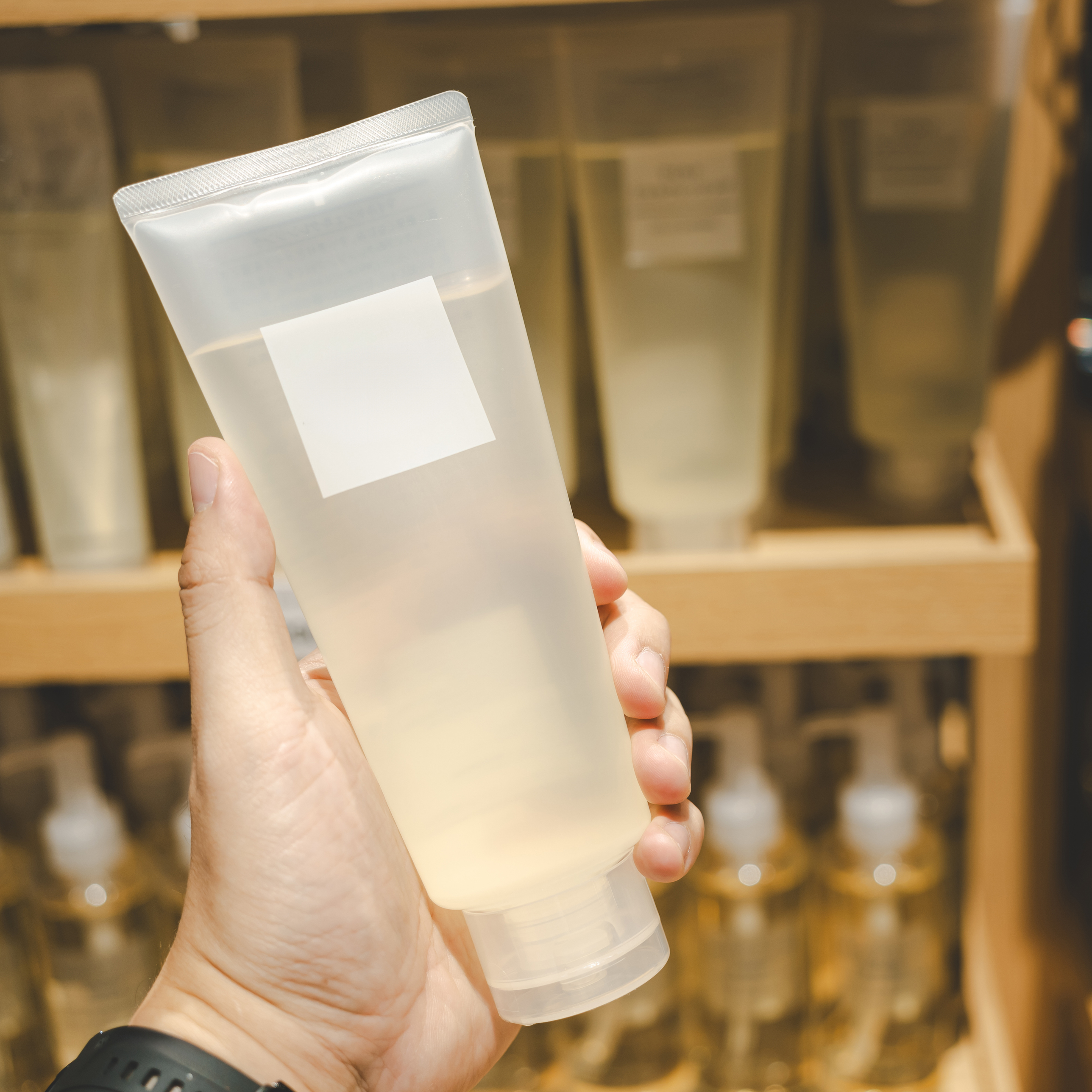 Eine Hand hält eine transparente Tube mit einer unbeschrifteten Etikette, umgeben von weiteren ähnlichen Produkten im Hintergrund.