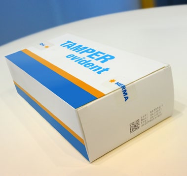 Verpackung mit dem Aufdruck "TAMPER evident" in Blau und Orange, auf einem Tisch platziert.