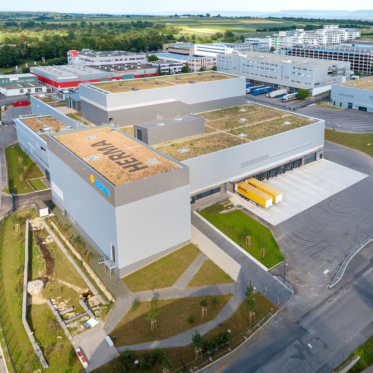 Moderne Lagerhalle mit begrüntem Dach, umgeben von Industriegebäuden und einer Straße. Sicht auf die Zufahrt und Lieferzone.