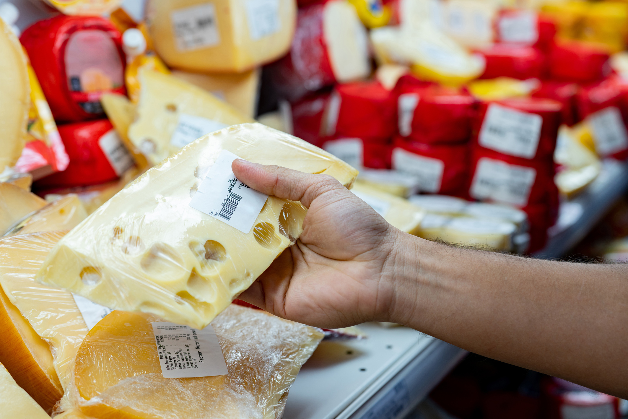 Eine Hand hält ein Stück Käse mit Löchern, während verschiedene Käsesorten im Hintergrund im Regal stehen.
