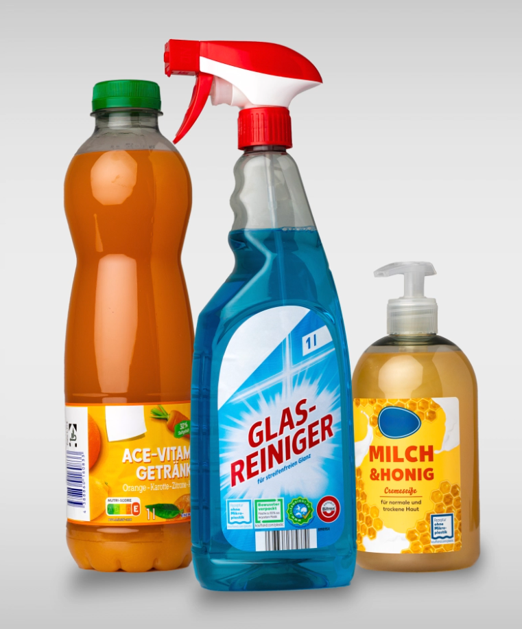 Drei Flaschen stehen nebeneinander: eine orange Getränkeflasche, eine blaue Glasreiniger-Sprühflasche und eine Flasche mit Milch- und Honigseife.