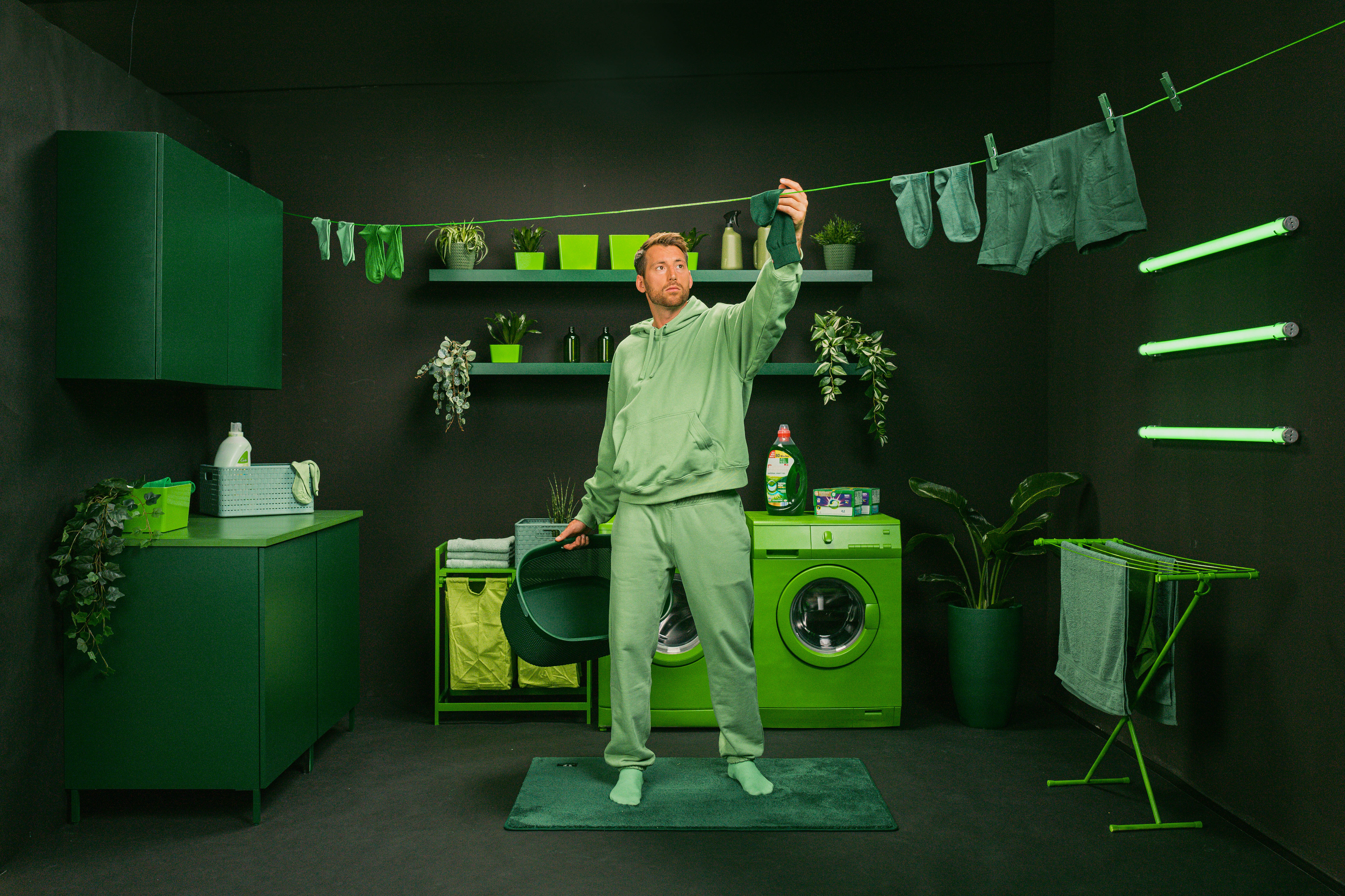Mann in grüner Kleidung hängt Wäsche in einem monochromen, grünen Raum auf. Waschmaschine und Pflanzen sind im Hintergrund sichtbar.