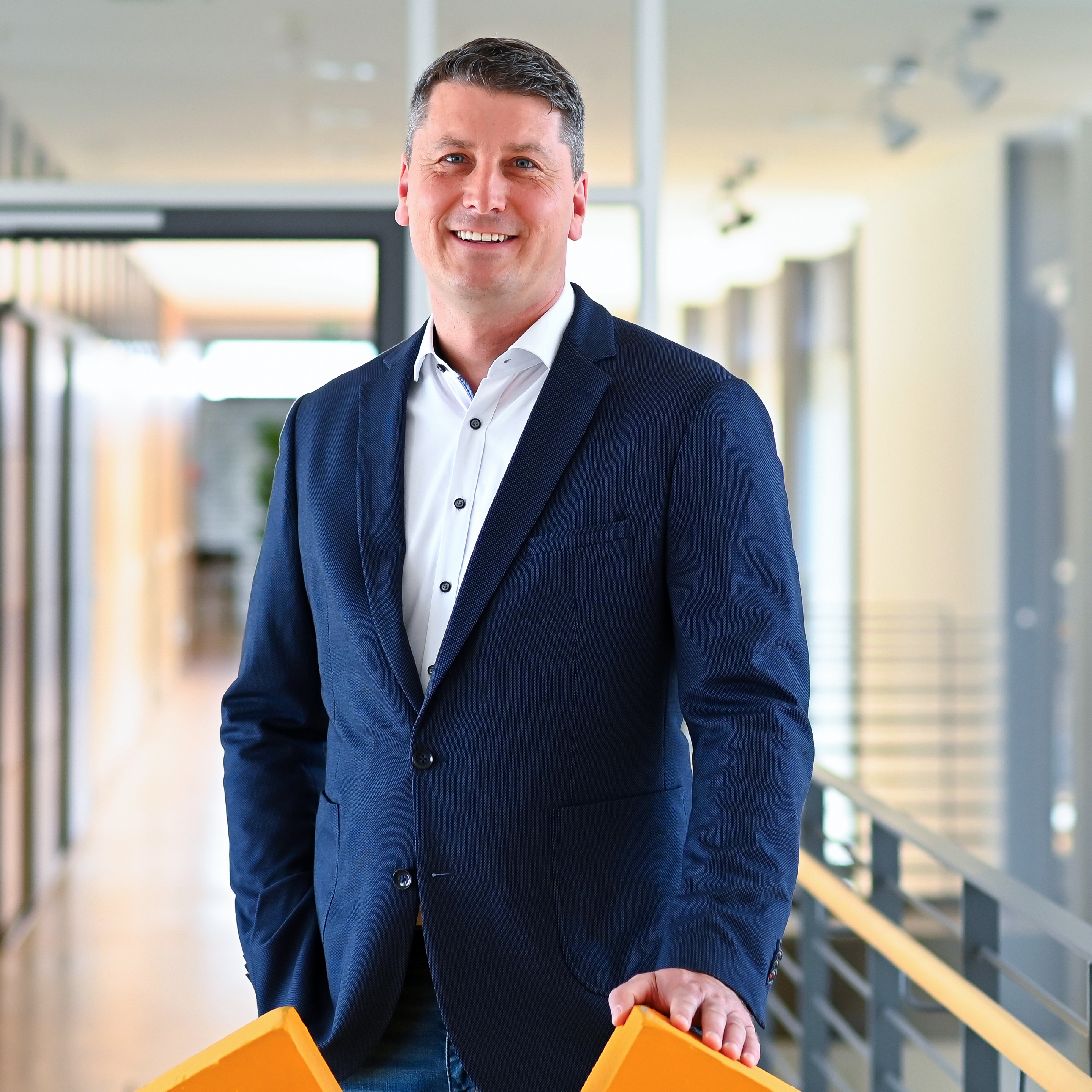 Ein Mann in einem blauen Blazer steht in einem modernen Büroflur und hält zwei orangefarbene Objekte in der Hand.