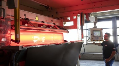 Ein Arbeiter steuert eine Maschine, die ein glühendes Material bearbeitet. Rote Beleuchtung und technische Geräte sind sichtbar.