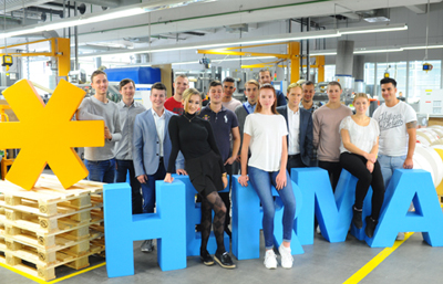 Eine Gruppe von jungen Menschen steht vor großen, bunten Buchstaben, die "HIMA" bilden, in einer modernen Produktionsumgebung.