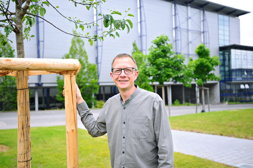 Mann mit Brille steht neben einem jungen Baum und einem Holzgestell vor einem modernen Gebäude mit großen Fenstern.