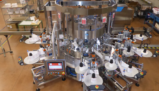Automatische Etikettiermaschine mit mehreren Stationen, die Produkte mit Etiketten versieht. Steuerungstafel im Vordergrund sichtbar.