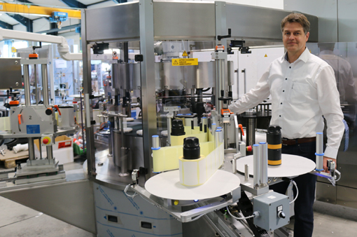 Ein Mann steht neben einer modernen Abfüll- und Etikettiermaschine in einer Produktionshalle, umgeben von Flaschen und Maschinen.