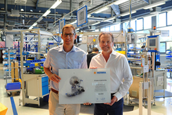 Zwei Männer halten ein Zertifikat in einer Produktionshalle, umgeben von Maschinen und Arbeitsplätzen.