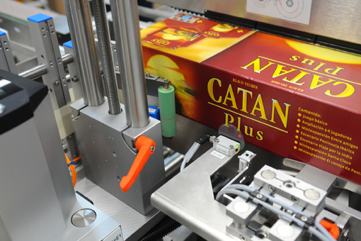 Eine Verpackungsmaschine, die eine Schachtel des Spiels "Catan Plus" verarbeitet, mit verschiedenen mechanischen Komponenten im Vordergrund.