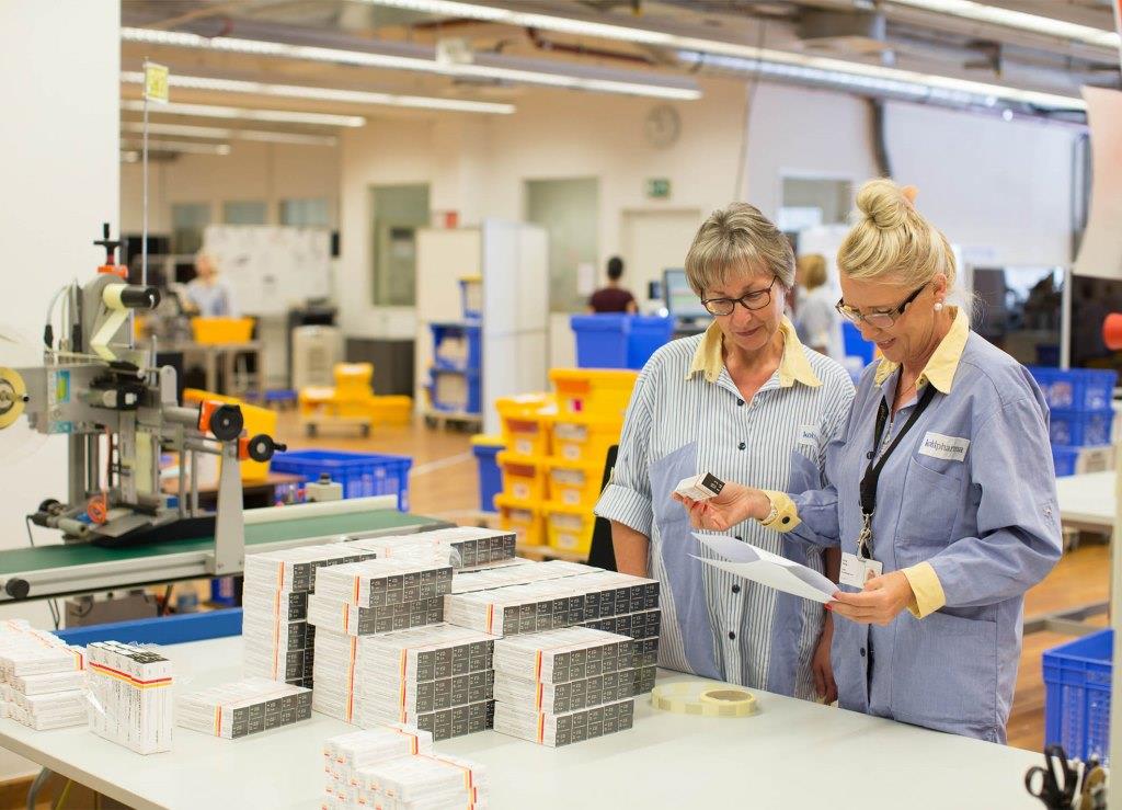 Zwei Frauen in Arbeitskleidung überprüfen Dokumente und Verpackungen an einem Tisch in einer Produktionsumgebung.