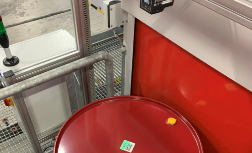 Roter Behälter mit einem Etikett und einem gelben Deckel, platziert in einer industriellen Umgebung mit Maschinen im Hintergrund.