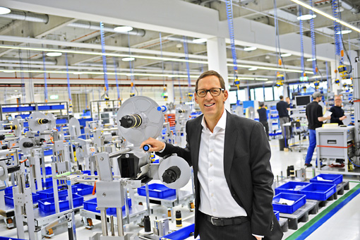 Ein Mann in einem Anzug steht in einer modernen Produktionshalle und hält ein Gerät. Im Hintergrund sind Arbeitsstationen und blaue Behälter sichtbar.
