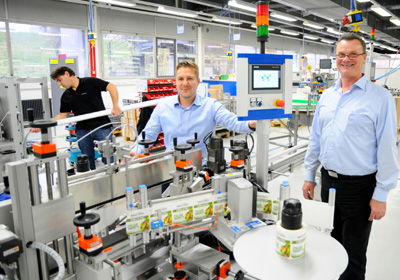 Zwei Männer stehen an einer Produktionslinie in einer Fabrik, während ein dritter Mitarbeiter im Hintergrund arbeitet.