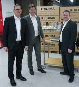 Vier Männer in Anzügen stehen vor einem Tisch mit verschiedenen Produkten und Kartons der Marke HERMA im Hintergrund.
